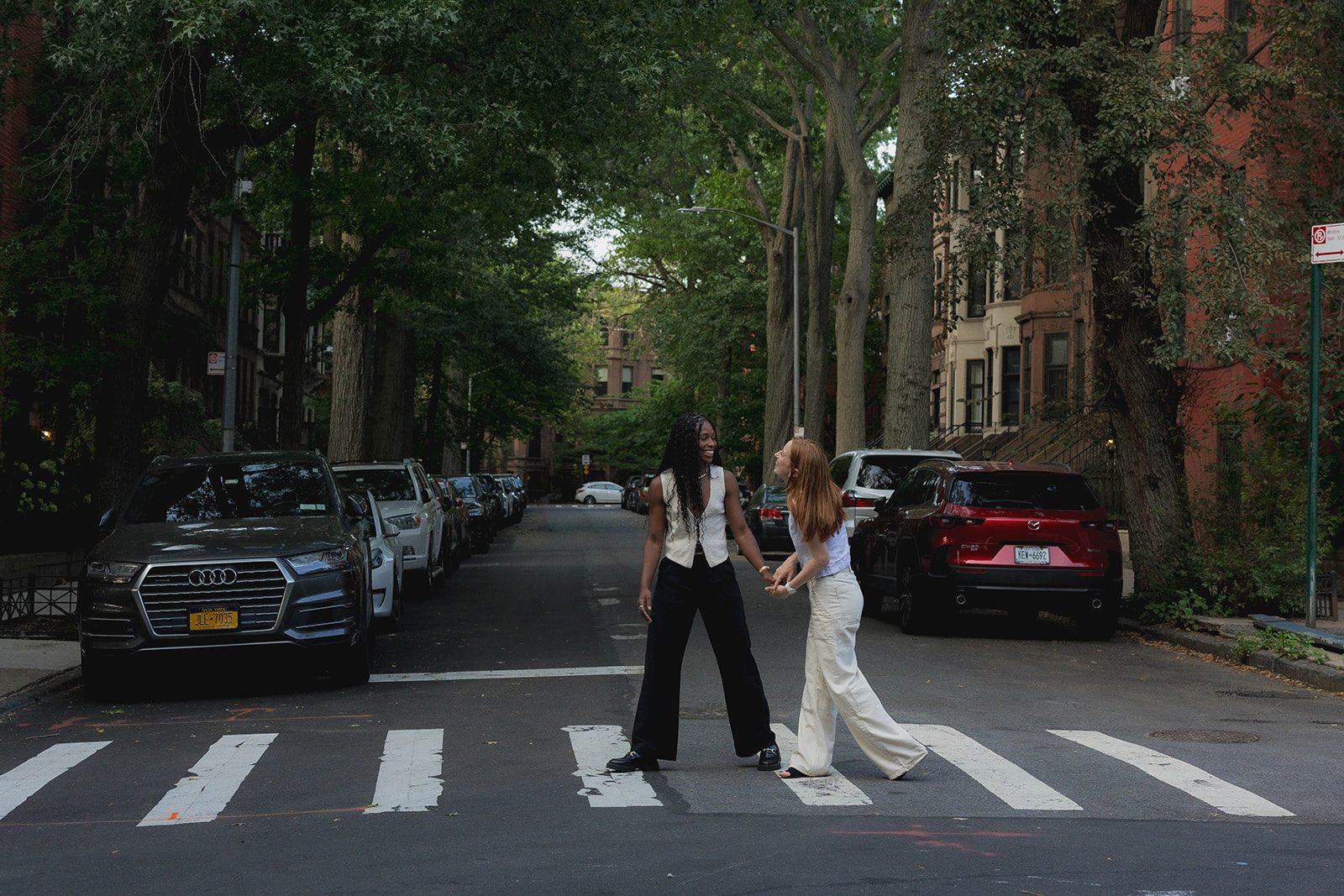 Cassia and Laura holding hands in a Park Slope crosswalk beneath a canopy of trees