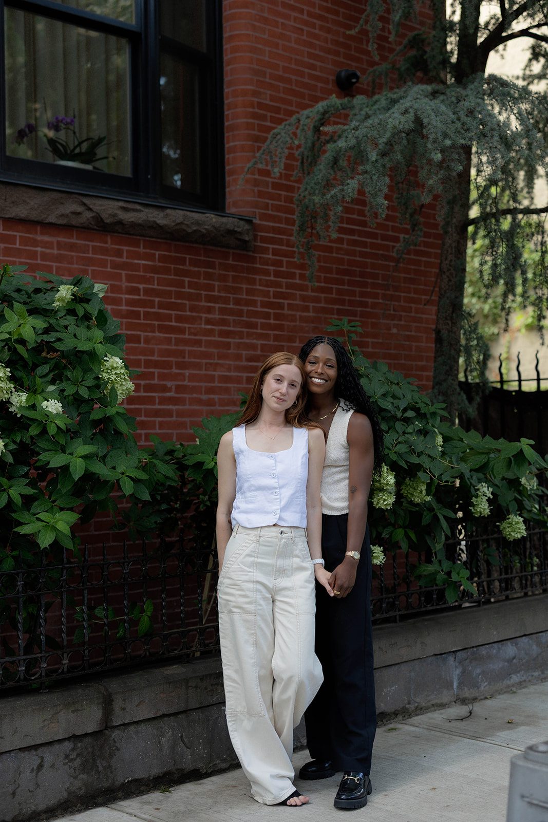 Cassia and Laura standing close together beside greenery and a red-brick Park Slope building